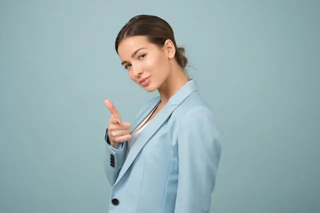 Confident woman in a light blue blazer smiling and pointing at the camera with a playful, self-assured gesture against a solid light blue background.